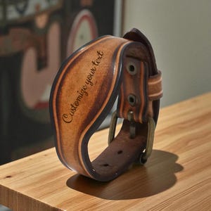 May include: A brown leather cuff bracelet with the text "Customize your text" engraved on the front. The bracelet has a buckle closure and metal hardware. The leather has a distressed look.