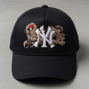 Puede incluir: Una gorra de béisbol negra con un logotipo NY bordado en blanco en la parte delantera. El logo está rodeado por un intrincado bordado de volutas de humo en tonos beige y marrón, con una pequeña figura de pelo rojo y un brazo esquelético.