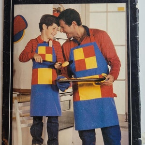 Puede incluir: Un patrón de costura vintage para un delantal a cuadros azules y rojos con un diseño de casa y árbol blancos. El patrón se titula "New Country Gear" y es de la empresa de patrones Butterick.