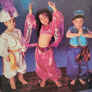 May include: Three children in Aladdin-themed costumes. One wears a white outfit with a gold sash and a feathered hat. Another wears a pink belly dancer outfit. The third child is in a blue and gold outfit with a matching hat.