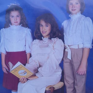 Puede incluir: Tres jóvenes posan para una foto con ropa vintage. Una persona está sentada, sosteniendo un libro titulado "The Tale of Squirrel Nutkin". Los demás están de pie, vistiendo blusas blancas con detalles de encaje y cuellos altos. El fondo es azul.