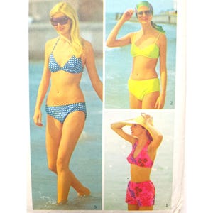 Könnte beinhalten: Eine Vintage-Modeillustration mit drei Frauen in Badebekleidung. Eine trägt einen blauen Bikini mit weißen Punkten, eine andere einen neongelben Bikini und die dritte einen rosa geblümten Bikini mit passenden Shorts. Alle posieren an einem Strand.