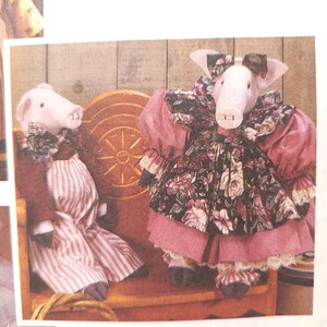 Op de afbeelding: Twee handgemaakte varkenspoppen gekleed in vintage-geïnspireerde kleding. Het varken links draagt een rode jurk met een witte schort en een roze strik. Het varken rechts draagt een roze jurk met een bloemenprint en een roze strik. Beide varkens zitten op een houten bank. De tekst "by/par Faye Wine" is onder de afbeelding gedrukt.