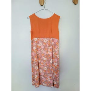 May include: Sleeveless orange dress with a floral print skirt. The top of the dress is a solid orange color, while the skirt features a pattern of pink, white, and orange flowers on a muted background. The dress is hanging on a metal hanger.