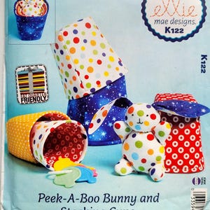 Könnte beinhalten: Ein Schnittmuster für Peek-A-Boo Bunny und Stacking Cups. Das Muster zeigt ein hasenförmiges Spielzeug und Stapelbecher. Die Becher haben bunte Punkte und Sternmuster. Der Text "ellie mae designs. K122" ist sichtbar.