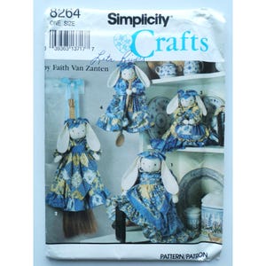 Può includere: Modello Simplicity Crafts 8264 per realizzare quattro figure di bambole a forma di coniglio. Le bambole sono vestite con tessuto a stampa floreale blu e giallo e hanno orecchie bianche. Il modello è di Faith Van Zanten.