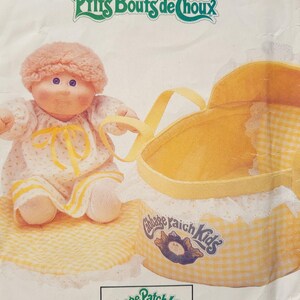 May include: A Cabbage Patch Kids doll wearing a yellow dress and a pink hat sits on a yellow and white checkered mat. The doll is holding a yellow and white checkered carrying case with a white lace trim. The carrying case has a Cabbage Patch Kids logo on the side. The image includes the text "Prits Bouts de Choux" and "Cabbage Patch Kids".