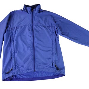 Könnte beinhalten: Blaue Jacke mit durchgehendem Reißverschluss, Stehkragen und langen Ärmeln. Die Jacke hat ein kleines Logo auf der Brust und zwei Seitentaschen mit Kordelzügen. Die Jacke ist aus leichtem Material.