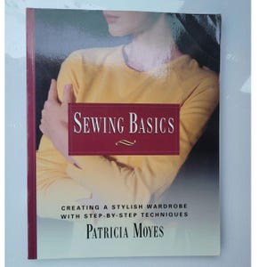 May include: A book titled "Sewing Basics" with a burgundy banner across the front. The banner has the title in white text. Below the title is the text "Creating a Stylish Wardrobe with Step-by-Step Techniques" and the author's name, Patricia Moyes.