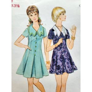 May include: Vintage sewing pattern showing two short-sleeved dresses. One is light blue with a scalloped collar and green trim, the other is purple with a white collar and floral pattern. The pattern includes measurements of 85 cm.