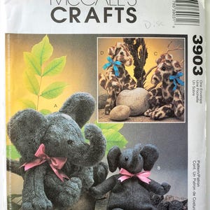 May include: McCall's Crafts pattern 3903 featuring stuffed animal designs. The image shows a gray elephant plush toy with a pink bow, and giraffe-like plush toys with blue bows. The pattern includes instructions for creating these soft toys.
