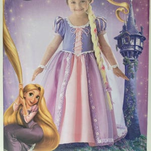 Può includere: Una busta per cartamodello Simplicity per un costume Disney Rapunzel, modello 2065, taglia A 3-8. La copertina mostra un abito viola e rosa con finiture in pizzo, una lunga parrucca bionda intrecciata e un'illustrazione di Rapunzel vicino a una torre.