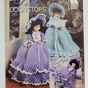 May include: A crochet pattern book titled "Dolly Doorstops" showing two doll doorstops in purple and blue dresses with matching hats. The cover also shows a smaller image of a doll, and the text "Makes A Great Pajama Bag Too!"