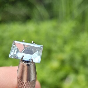 Può includere: Una pietra preziosa rettangolare sfaccettata di colore azzurro, tenuta in una montatura in metallo a griffe. La pietra è trasparente con bordi netti e una superficie lucida che riflette la luce sulla parte superiore piatta e sui lati angolati.
