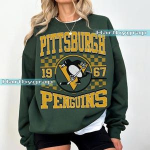 Pu&ograve; includere: Felpa verde scuro con la scritta "PITTSBURGH PENGUINS 1967" in oro. Il design include un logo di pinguino con una mazza da hockey e un triangolo giallo. Girocollo, maniche lunghe.
