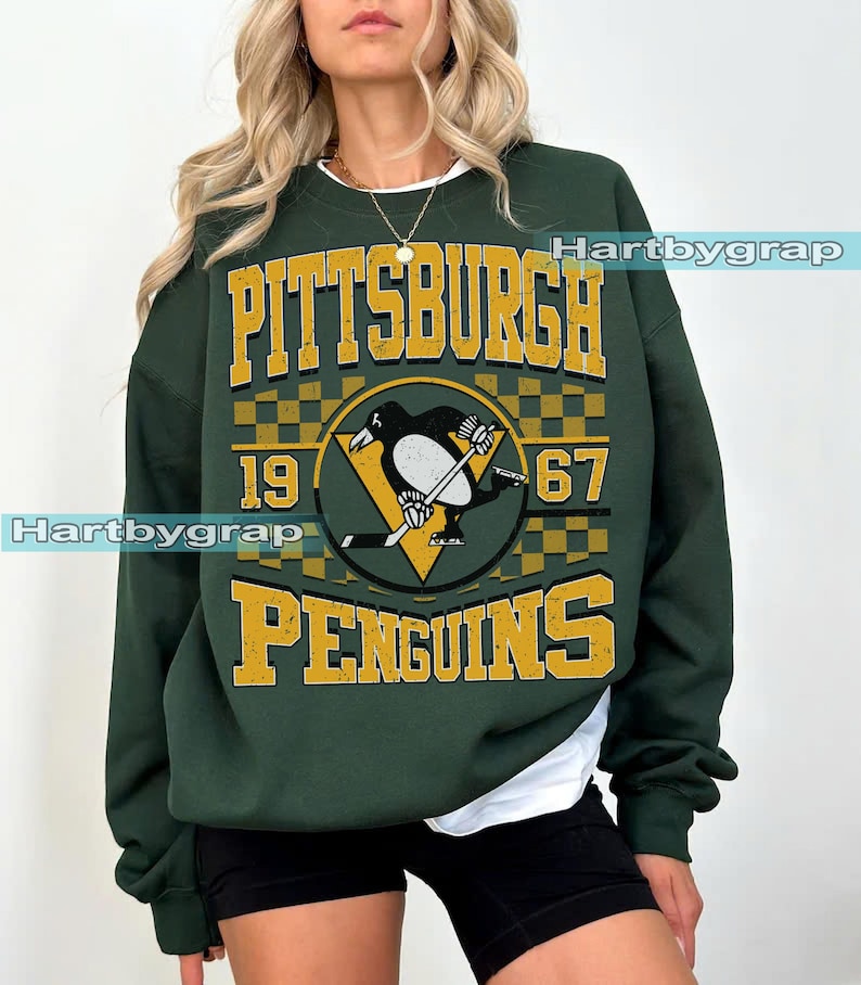Pu&ograve; includere: Felpa verde scuro con la scritta "PITTSBURGH PENGUINS 1967" in oro. Il design include un logo di pinguino con una mazza da hockey e un triangolo giallo. Girocollo, maniche lunghe.