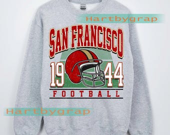 May include: A grey sweatshirt with a vintage San Francisco football design. The design includes a red football helmet, the words "SAN FRANCISCO" in red, and the year "1944". The word "FOOTBALL" is printed below the year.