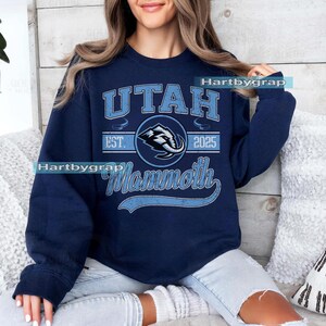 May include: Navy blue sweatshirt with a light blue graphic that reads "UTAH Mammoth 2025". The graphic includes a stylized mammoth logo. The sweatshirt is a crew neck style.