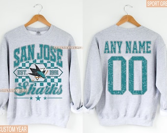 May include: Grey sweatshirt with the San Jose Sharks logo in teal and white. The front features the team name and logo, and the back has space for a custom name and number. The sweatshirt is displayed on a wooden hanger.