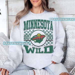 Op de afbeelding: Een grijze sweatshirt met de tekst "MINNESOTA WILD" in groen en wit, en een cirkelvormig logo van een hockeyteam. Het logo toont een boslandschap en de cijfers "20 00".