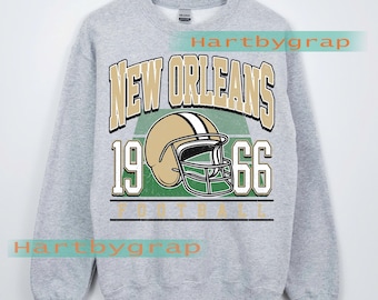 May include: A gray sweatshirt with a vintage-style design. The design features the words "NEW ORLEANS" in gold lettering above a gold football helmet. Below the helmet are the numbers "1966" in white. The word "FOOTBALL" is printed below the numbers.