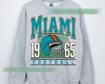 Könnte beinhalten: Ein graues Sweatshirt mit einer türkis-orangenen Grafik eines Football-Helms und dem Wort "MIAMI" über den Zahlen "1965". Das Wort "FOOTBALL" befindet sich unter dem Helm. Die Worte "CUSTOM YEAR" stehen unten.