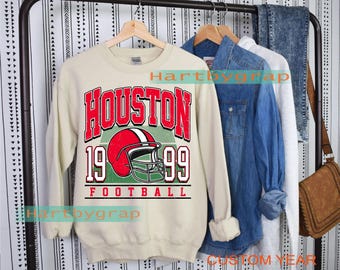 Op de afbeelding: Crèmekleurige sweatshirt met een rood en wit "HOUSTON 1999 FOOTBALL"-opdruk en een voetbalhelm-ontwerp. De sweatshirt hangt aan een kledingrek met andere kledingstukken, waaronder spijkeroverhemden en jeans.