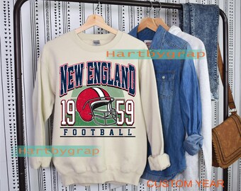 May include: Cream-colored sweatshirt with a graphic design featuring "NEW ENGLAND" in navy blue, a red and white football helmet, and the numbers "1959." The word "FOOTBALL" is printed below. Other clothing items hang in the background.