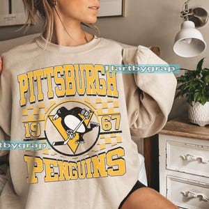 Pu&ograve; includere: Felpa beige con la scritta "PITTSBURGH PENGUINS" in lettere gialle. Il design include un logo di pinguino nero con una mazza da hockey e i numeri "1967". La felpa ha maniche lunghe.