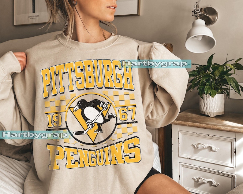 Pu&ograve; includere: Felpa beige con la scritta "PITTSBURGH PENGUINS" in lettere gialle. Il design include un logo di pinguino nero con una mazza da hockey e i numeri "1967". La felpa ha maniche lunghe.