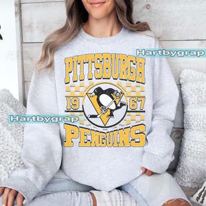 Pu&ograve; includere: Una felpa grigio chiaro con la scritta "PITTSBURGH PENGUINS" in lettere gialle. Il design include un logo di pinguino con una mazza da hockey e i numeri "1967". La felpa &egrave; in stile casual.