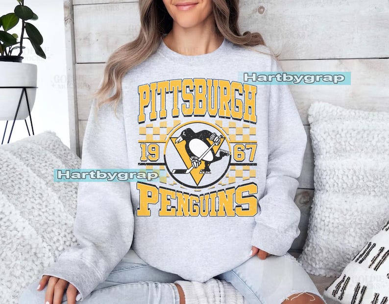 Pu&ograve; includere: Una felpa grigio chiaro con la scritta "PITTSBURGH PENGUINS" in lettere gialle. Il design include un logo di pinguino con una mazza da hockey e i numeri "1967". La felpa &egrave; in stile casual.