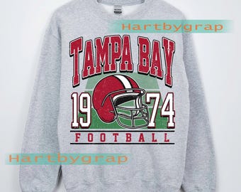 Puede incluir: Una sudadera gris con un gráfico desgastado que presenta "TAMPA BAY" en letras rojas, un casco de fútbol americano rojo, el número "1974" y la palabra "FOOTBALL". El texto "CUSTOM YEAR" está en la parte inferior.