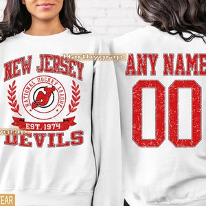 May include: White sweatshirt featuring the New Jersey Devils logo and text. The front displays "NEW JERSEY" above a circular logo with "NATIONAL HOCKEY LEAGUE" and "EST. 1974". The back has "ANY NAME" and the number "00" in red.