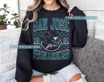 May include: A black sweatshirt with the words "SAN JOSE SHARKS" in teal and white, and a shark logo. The year "1991" is also visible. The sweatshirt is being worn.