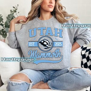 May include: A gray sweatshirt with the text "UTAH Mammoth" in light blue. The design includes a mammoth logo inside a circle, with "EST. 2025" above. The sweatshirt is worn by a person wearing blue jeans.