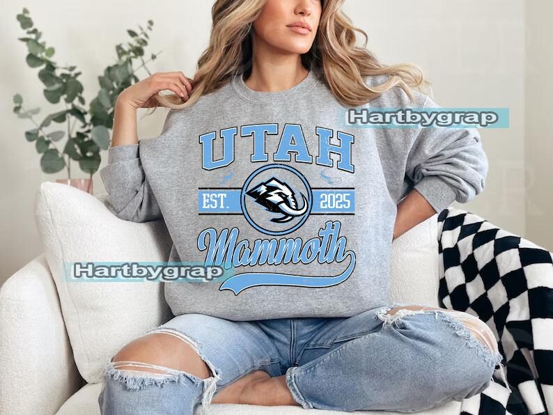 May include: A gray sweatshirt with the text "UTAH Mammoth" in light blue. The design includes a mammoth logo inside a circle, with "EST. 2025" above. The sweatshirt is worn by a person wearing blue jeans.