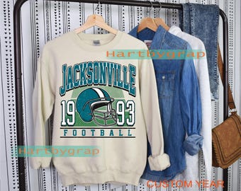 May include: A cream-coloured sweatshirt featuring "JACKSONVILLE 1993 FOOTBALL" text and a teal football helmet graphic. The sweatshirt is hanging on a clothes rail with denim and other shirts.