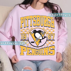 Pu&ograve; includere: Una felpa rosa chiaro con la scritta "PITTSBURGH PENGUINS" in lettere gialle. Il design include un logo di pinguino in bianco e nero con una mazza da hockey e i numeri "1967".