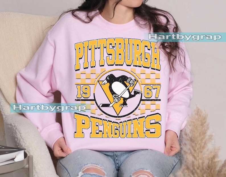 Pu&ograve; includere: Una felpa rosa chiaro con la scritta "PITTSBURGH PENGUINS" in lettere gialle. Il design include un logo di pinguino in bianco e nero con una mazza da hockey e i numeri "1967".