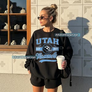 May include: Black sweatshirt with the text "UTAH Mammoth" in light blue. The sweatshirt has a crew neck and is paired with black shorts. The image also includes a coffee cup and sunglasses.