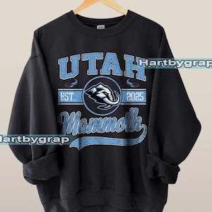 May include: Black crewneck sweatshirt with the text "UTAH Mammoth" in light blue. The design includes a circular emblem with an elephant graphic and the year "2025". The sweatshirt has a vintage, distressed look.