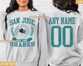 May include: Light grey sweatshirt with the San Jose Sharks logo and text reading "SAN JOSE SHARKS" and "EST. 1991". The back features "ANY NAME" and the number "00" in teal. A custom year option is available.