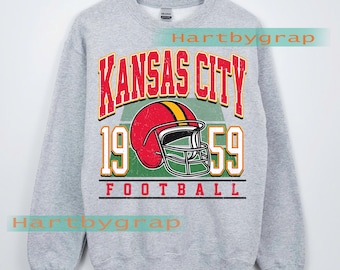 Könnte beinhalten: Ein hellgraues Sweatshirt mit einem Used-Look-Grafikdesign. Das Design zeigt die roten Worte "Kansas City", das Jahr "1959" und einen roten Footballhelm. Das Wort "FOOTBALL" ist darunter gedruckt.