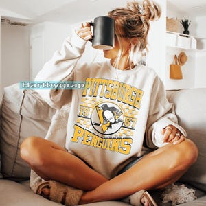 Pu&ograve; includere: Felpa beige con la scritta "PITTSBURGH PENGUINS" in giallo e nero, e un logo a forma di pinguino. Una persona tiene in mano una tazza nera. L'immagine &egrave; stata scattata in interni.