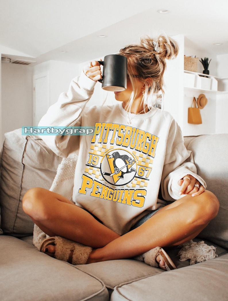 Pu&ograve; includere: Felpa beige con la scritta "PITTSBURGH PENGUINS" in giallo e nero, e un logo a forma di pinguino. Una persona tiene in mano una tazza nera. L'immagine &egrave; stata scattata in interni.