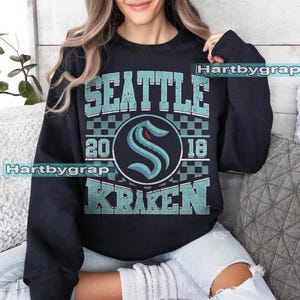 Pode incluir: Camisola preta com o texto "SEATTLE KRAKEN" em letras turquesas. O design inclui o logotipo Kraken e os números "20" e "18". A camisola é de estilo gola redonda.