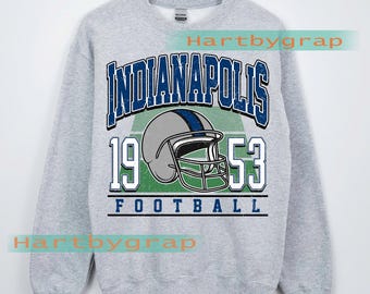 May include: A light grey sweatshirt with a vintage Indianapolis football design. The design features the word "Indianapolis" in blue, a football helmet, the numbers "1953", and the word "Football". The words "Custom Year" are at the bottom.