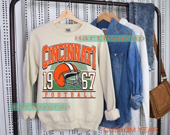 Può includere: Felpa color crema con la scritta "CINCINNATI 1967 FOOTBALL" in arancione e bianco. Il design include un casco da football americano arancione su sfondo verde. Altri capi d'abbigliamento sono appesi a un attaccapanni dietro la felpa.