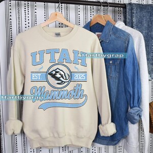 May include: Cream-colored sweatshirt with the text "UTAH Mammoth EST. 2025" in light blue. The design includes a circular emblem with a mammoth graphic. Other clothing items are visible in the background.